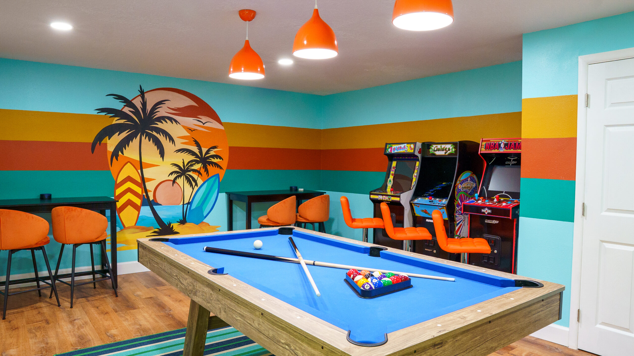 Garage conversion in Tampa, FL turned into a retro beach-themed game room with a hand-painted palm tree mural, striped walls, pool table, arcade machines, orange pendant lights, and wood-look vinyl flooring.