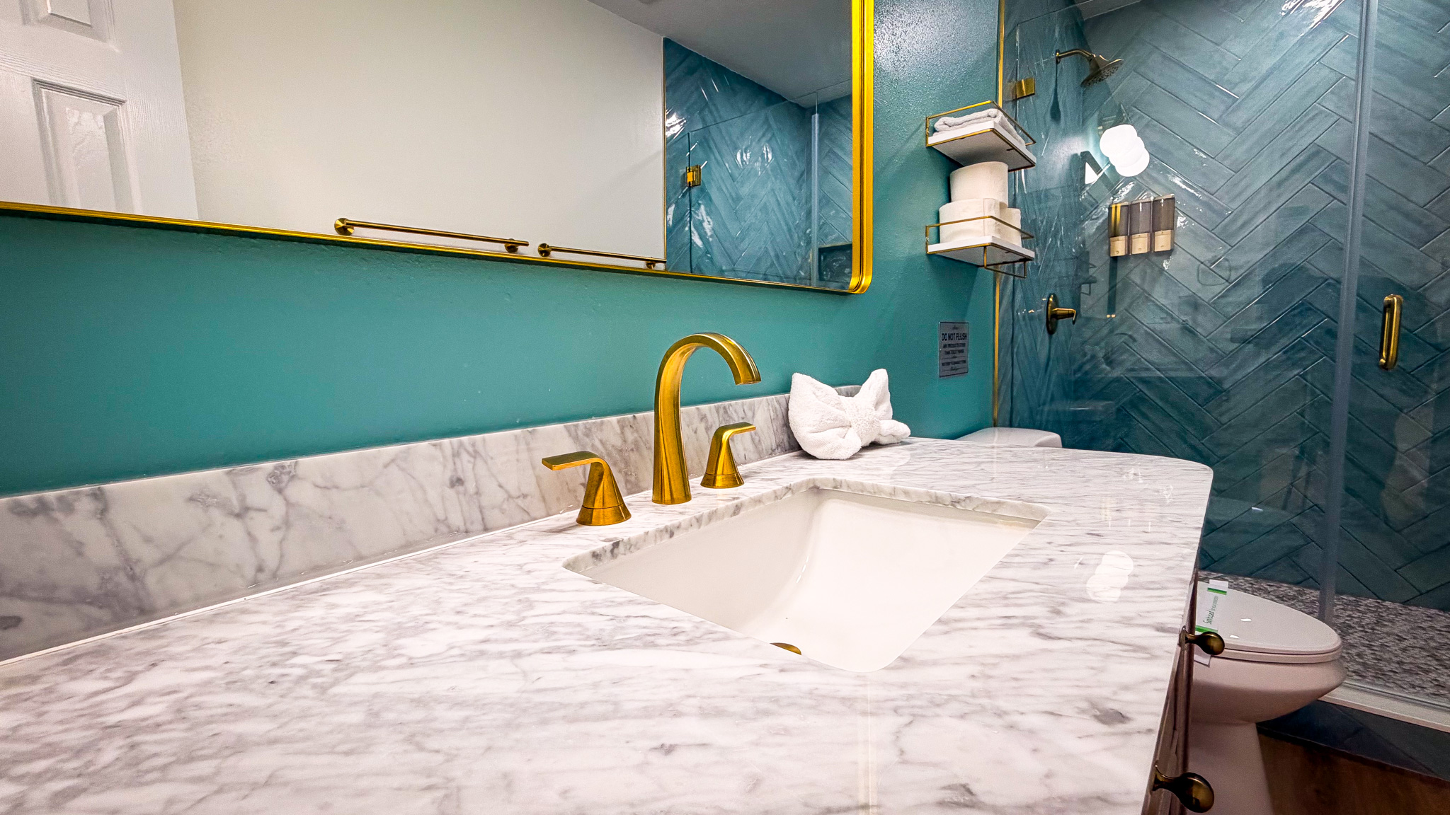 Bathroom remodel in Tampa, FL with a marble vanity, brushed gold faucet, and a glass walk-in shower with teal herringbone tile.