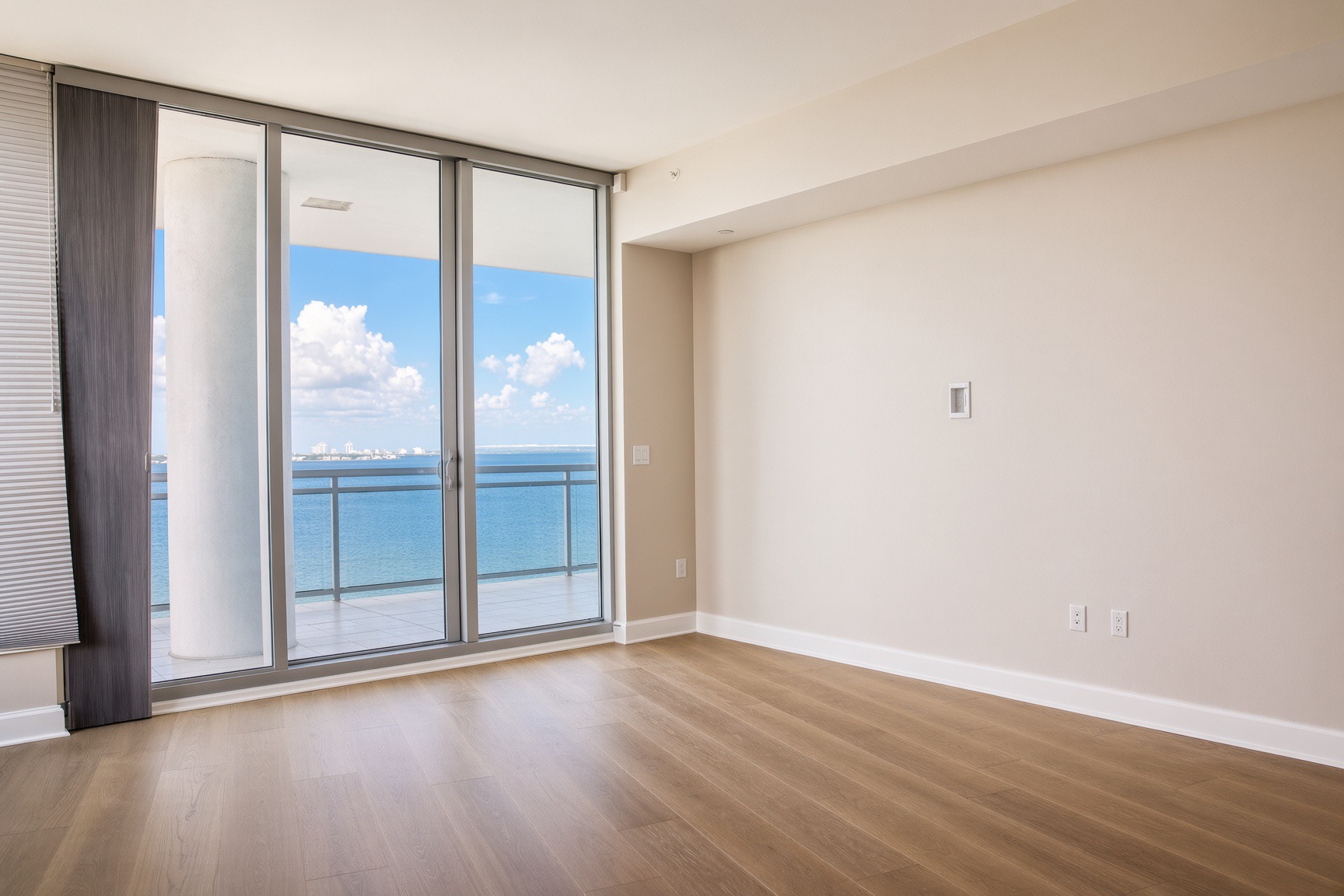 Renovated high-rise condo bedroom in Tampa, FL with warm wood-look flooring, new baseboards, neutral paint, and sliding glass doors to a waterfront balcony.
